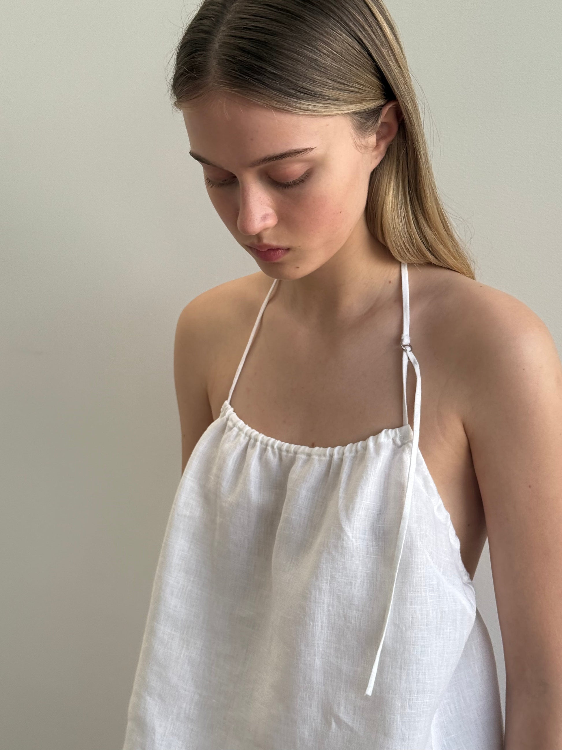 A young woman with long, straight hair looks down while wearing the O Ring Halter - White from Deiji Studios against a plain light-colored background.