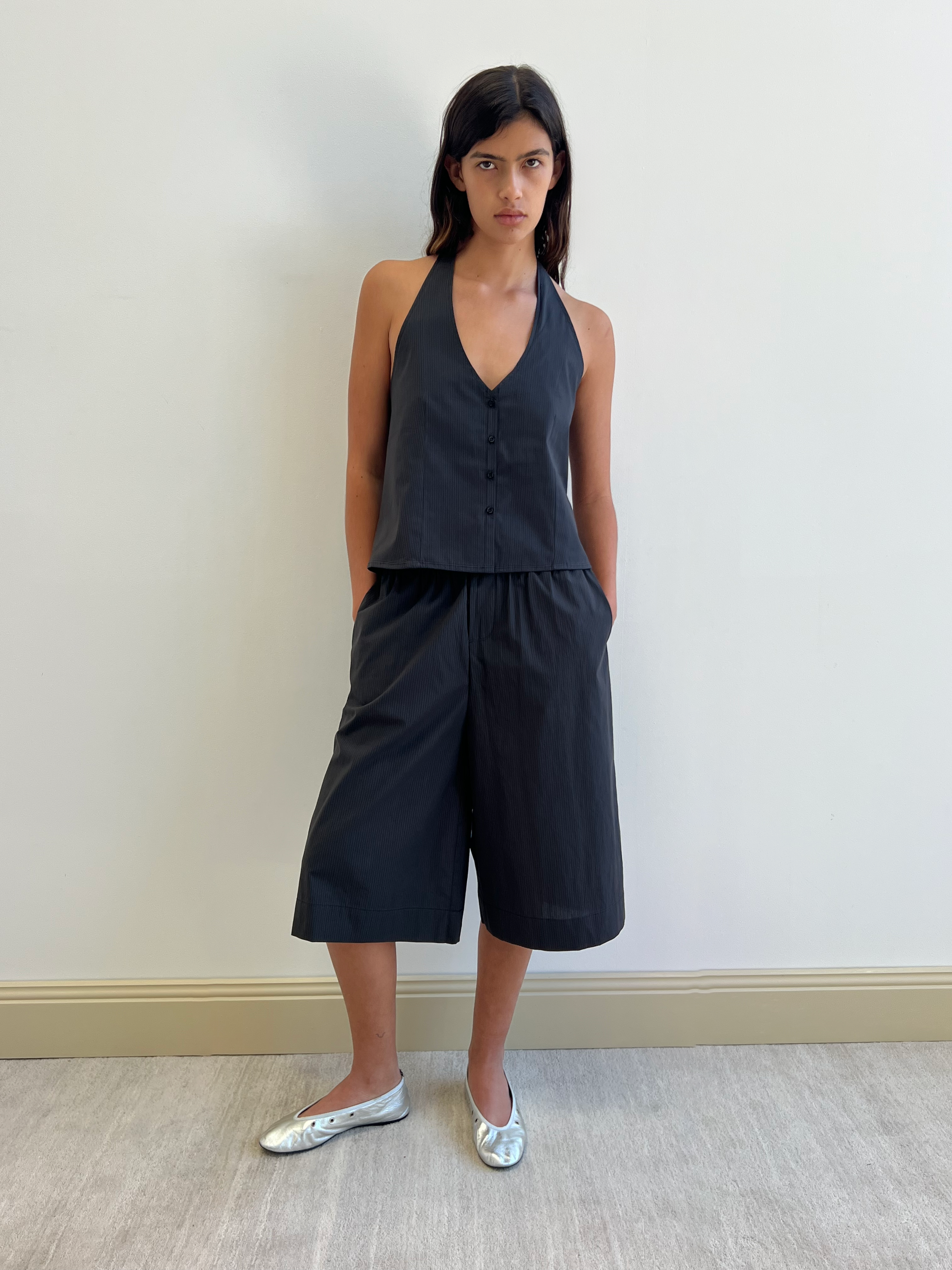 A woman wears the Long Lounger - Black Stripe from Deiji Studios, featuring a sleeveless, deep V-neck top with matching wide-leg shorts. She stands against a white wall with her hands in her pockets, paired with shiny silver flats.