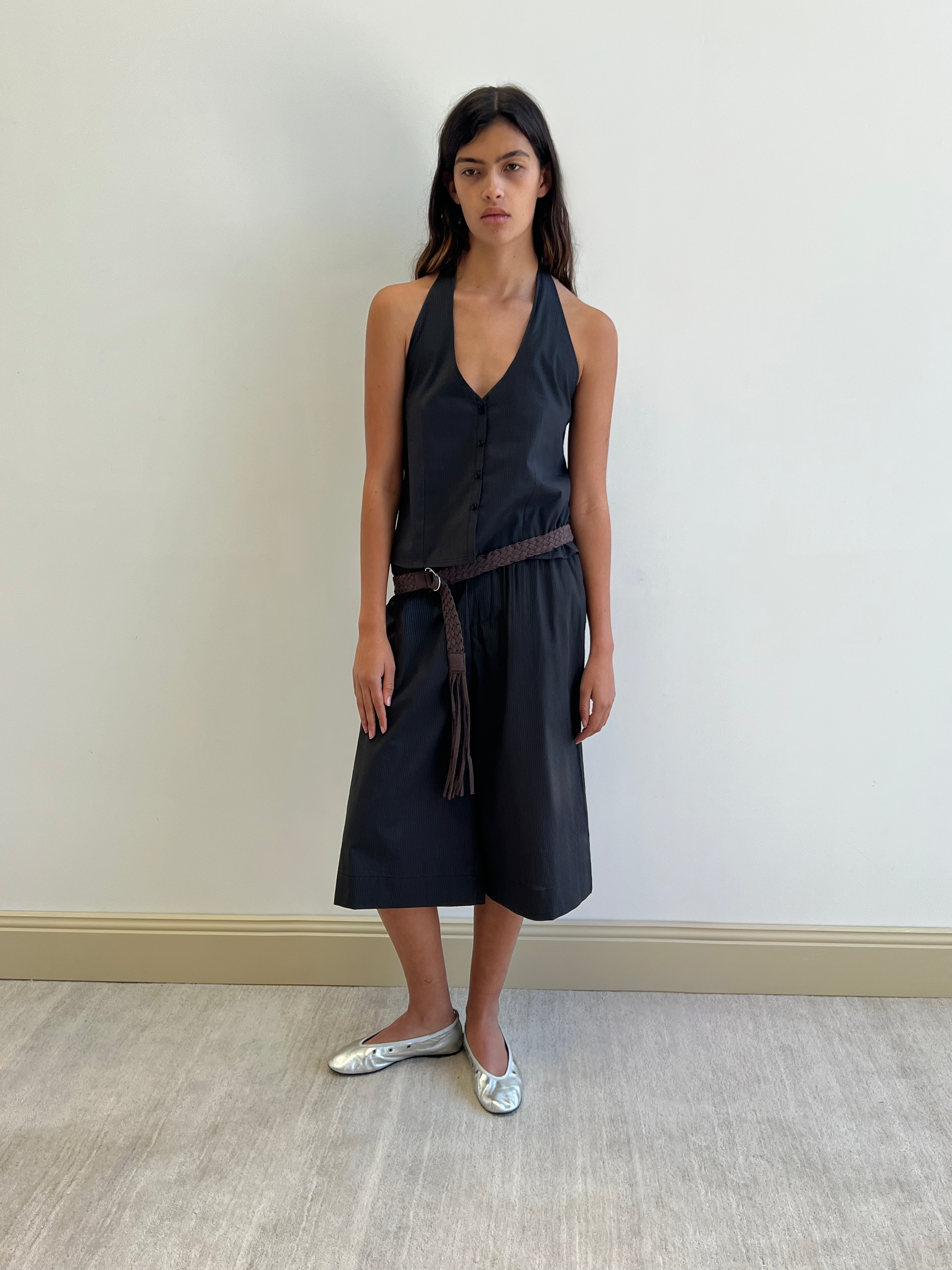 A woman with long dark hair and a neutral expression stands against a plain white wall, wearing the Long Lounger in Black Stripe—an organic cotton, sleeveless black halter dress—styled with a brown braided belt and silver ballet flats.
