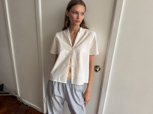 A woman stands indoors against a white wall, wearing the Sails Top - White made of organic cotton with loose buttons and light blue striped pants. She gazes thoughtfully to the side with one hand in her pocket.