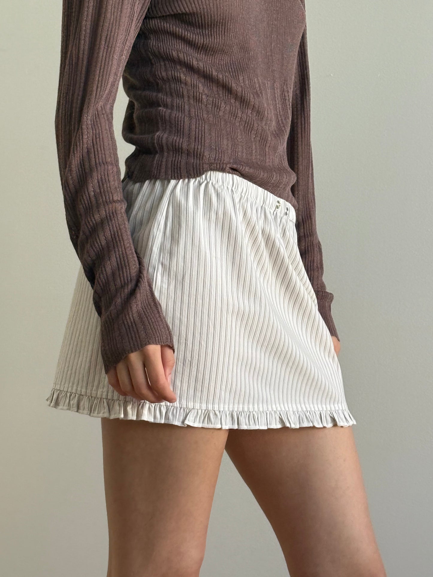 Wearing the Flounce Mini - Gull Stripe skirt by Deiji Studios and a ribbed, long-sleeve brown top, a person stands against a plain light background. The photo is cropped from the shoulders down.