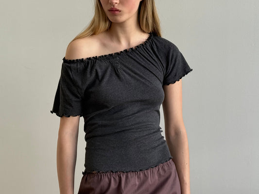 A woman wears the Flounce Tee - Grey by Deiji Studios with a mauve skirt, standing against a plain light grey background. Her hair is down and her face is partially out of frame.