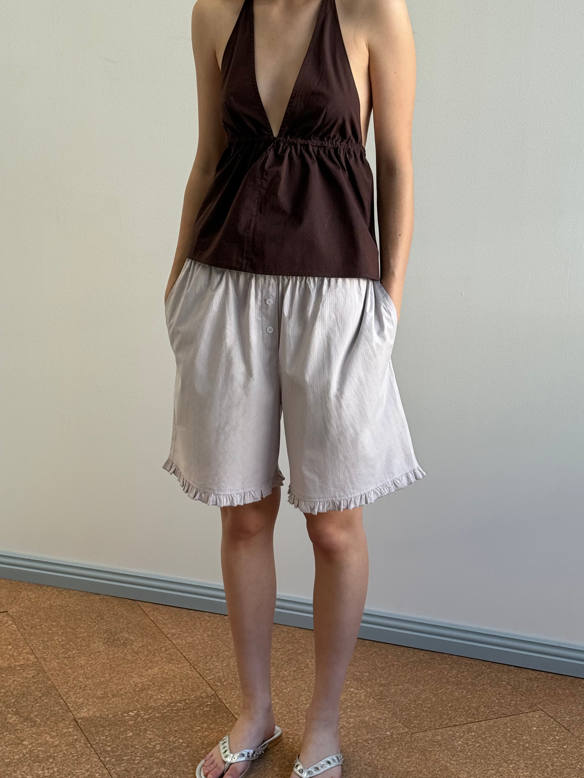 A person stands indoors wearing a sleeveless dark brown halter top and Deiji Studios' Flounce Short in Lilac Stripe, hands in pockets with white sandals, posing against a plain background with a blue baseboard.