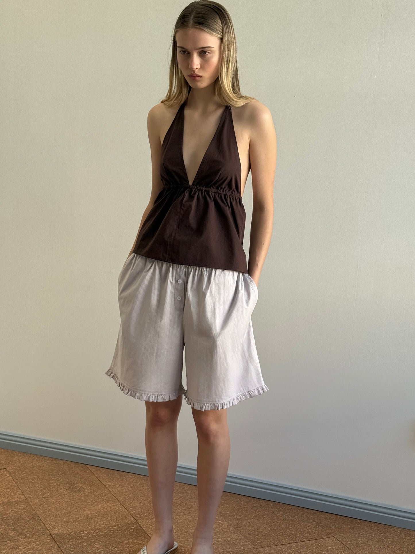 A woman with straight light brown hair and a neutral expression stands indoors against a plain wall, wearing a sleeveless dark brown halter top and relaxed fit Flounce Short - Lilac Stripe featuring ruffled hems.