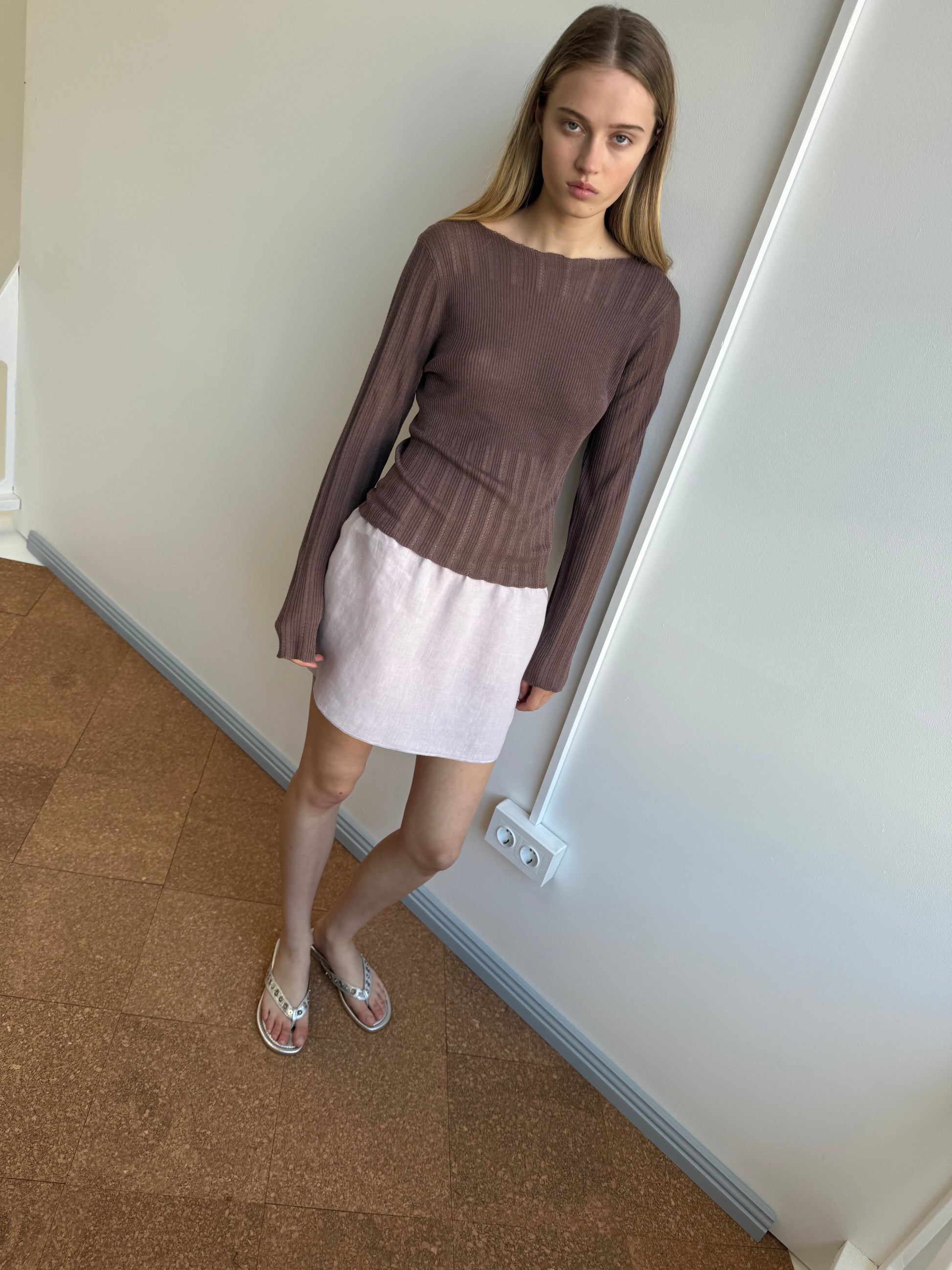 A young woman with straight blonde hair stands against a white wall, wearing a sheer brown ribbed long-sleeve top, silver embellished flip-flops, and the Babylock Skirt in lilac by Deiji Studios.