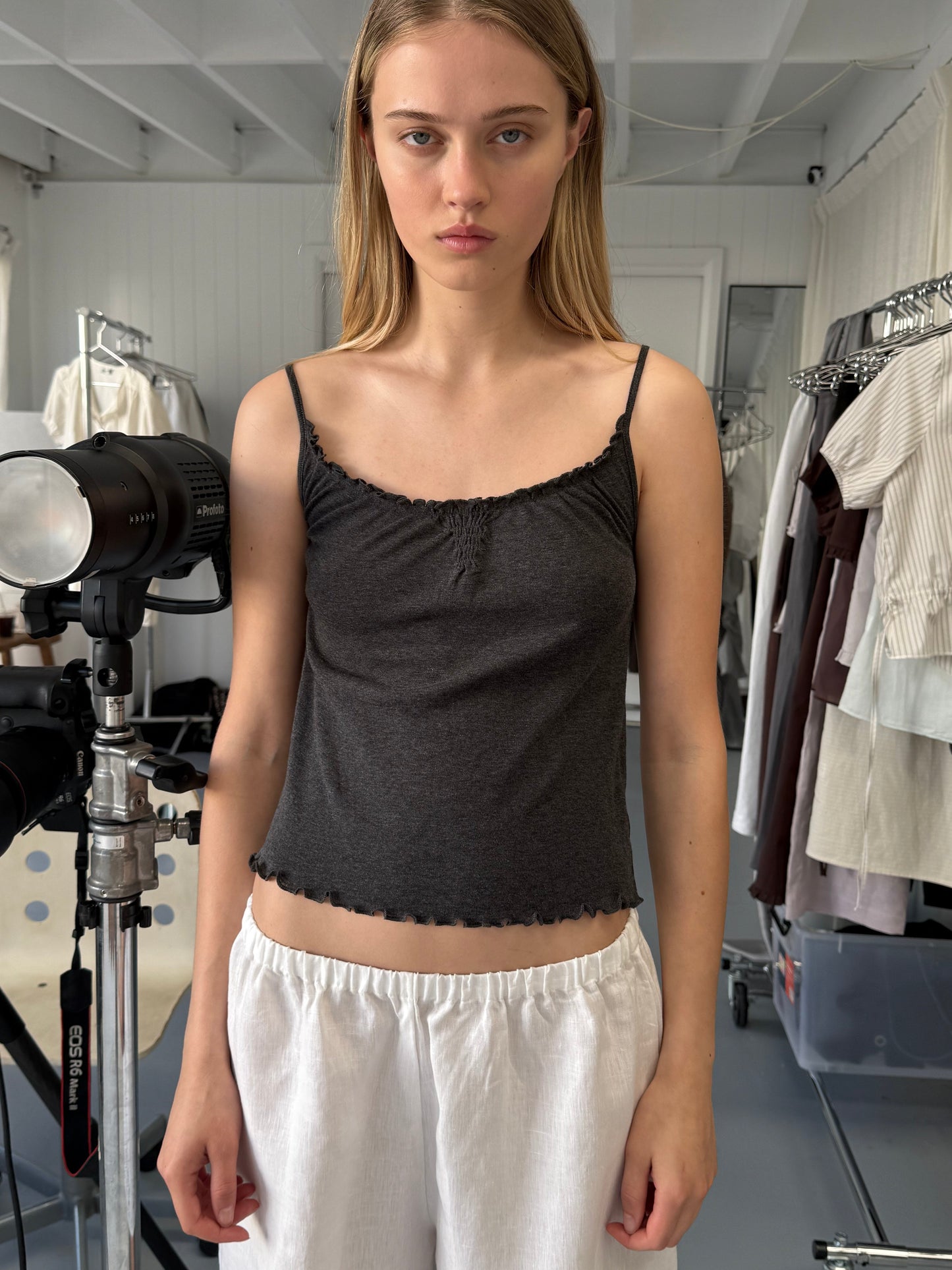 A woman with long blonde hair wears the Wool Blend Flounce Tank - Grey by Deiji Studios and white pants, standing indoors in a studio with lighting equipment and clothing racks, looking at the camera with a neutral expression.
