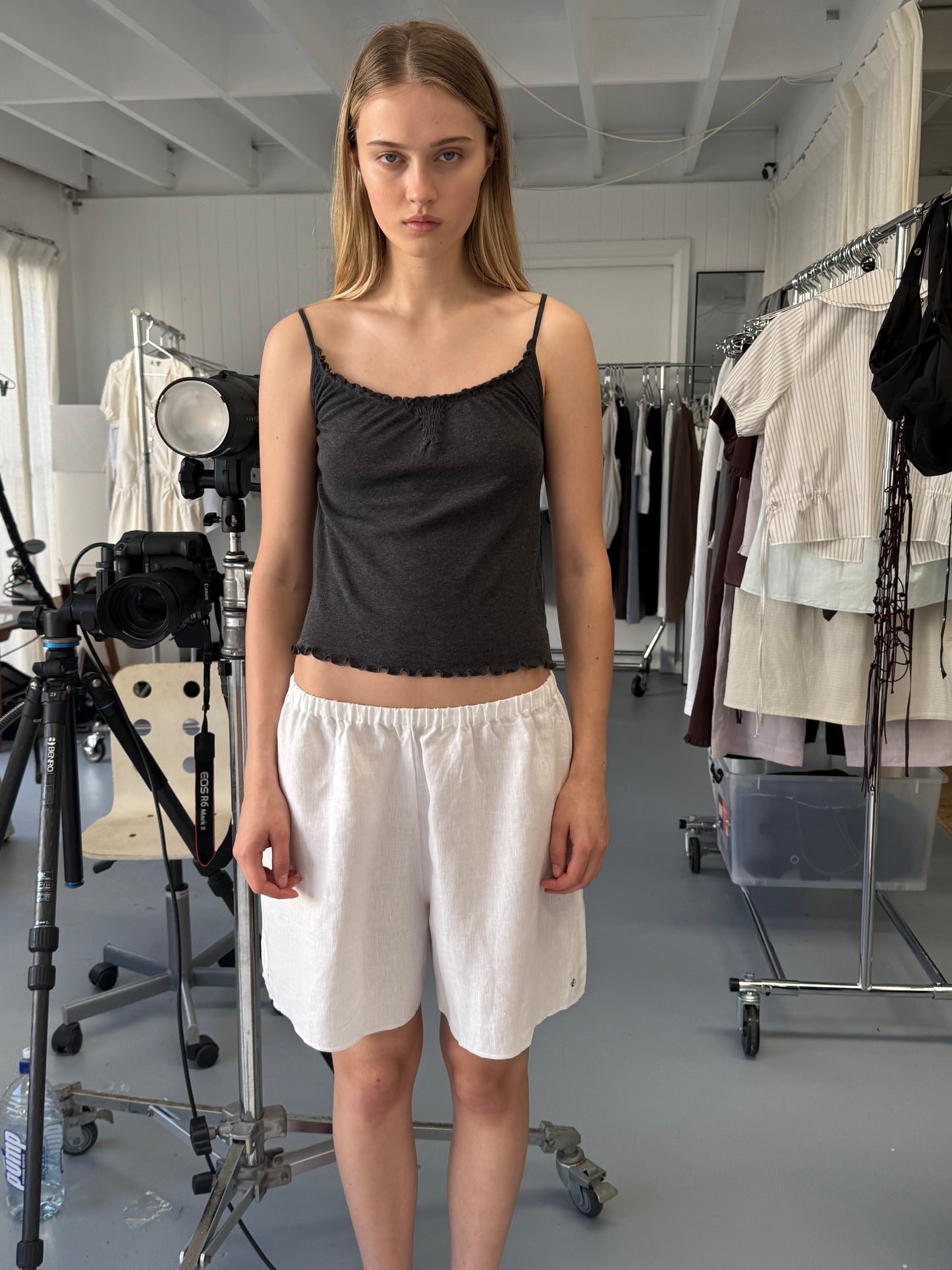 A young woman with straight blonde hair stands indoors wearing a black tank top and the Press Stud Short. She's surrounded by clothing racks, studio lights, and a camera tripod in a well-lit room.