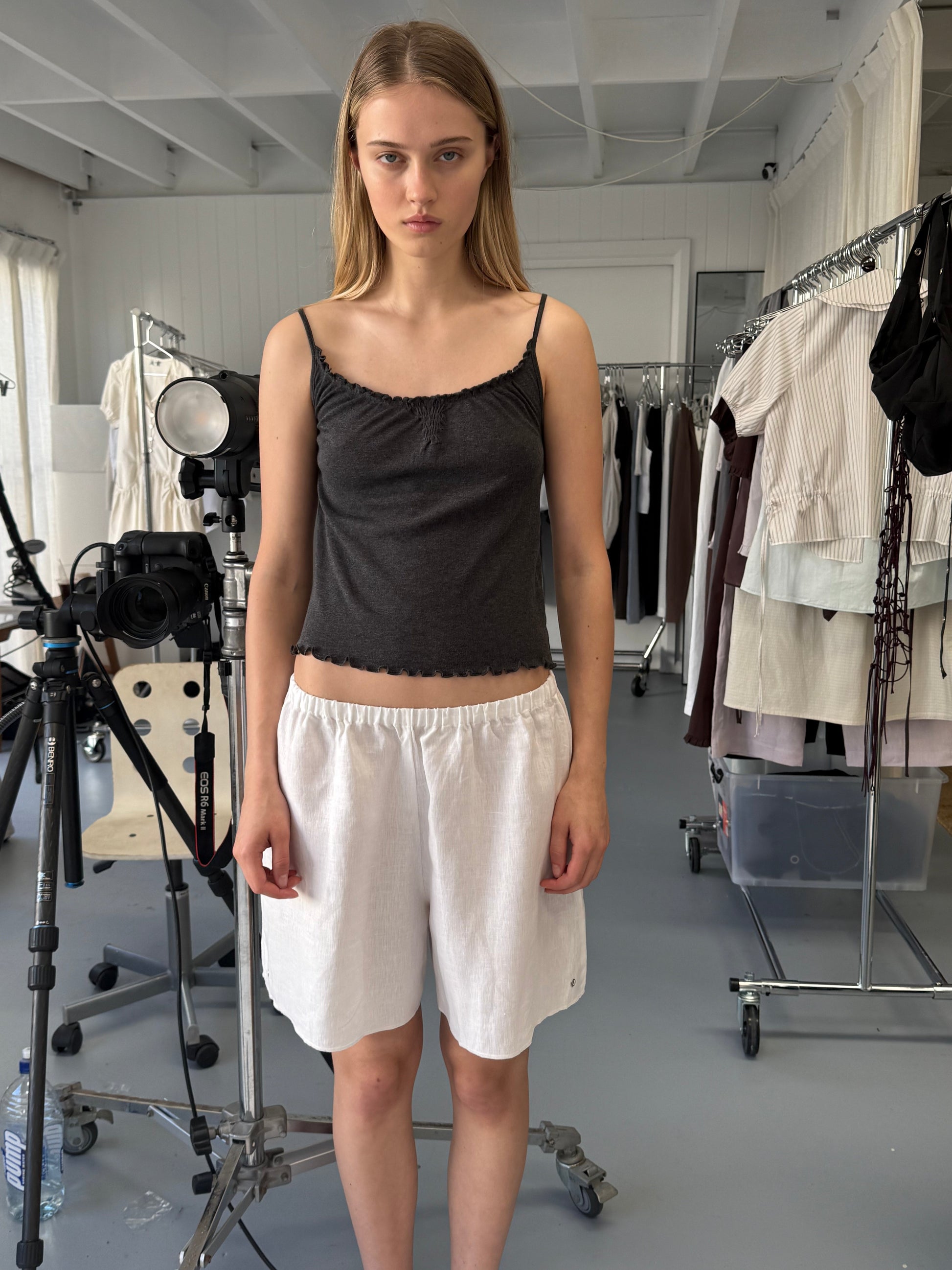A young woman with straight blonde hair stands indoors wearing a black tank top and the Press Stud Short. She's surrounded by clothing racks, studio lights, and a camera tripod in a well-lit room.