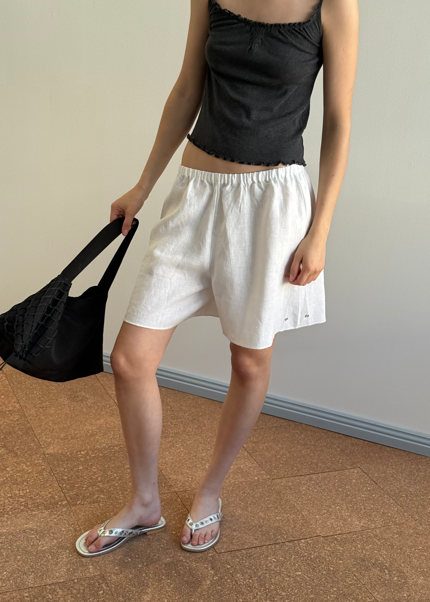 A person stands indoors in a black sleeveless top, white Deiji Studios Press Stud Shorts with an elastic waist, white sandals, and holding a black woven bag. The background features a tiled floor and light-colored wall.
