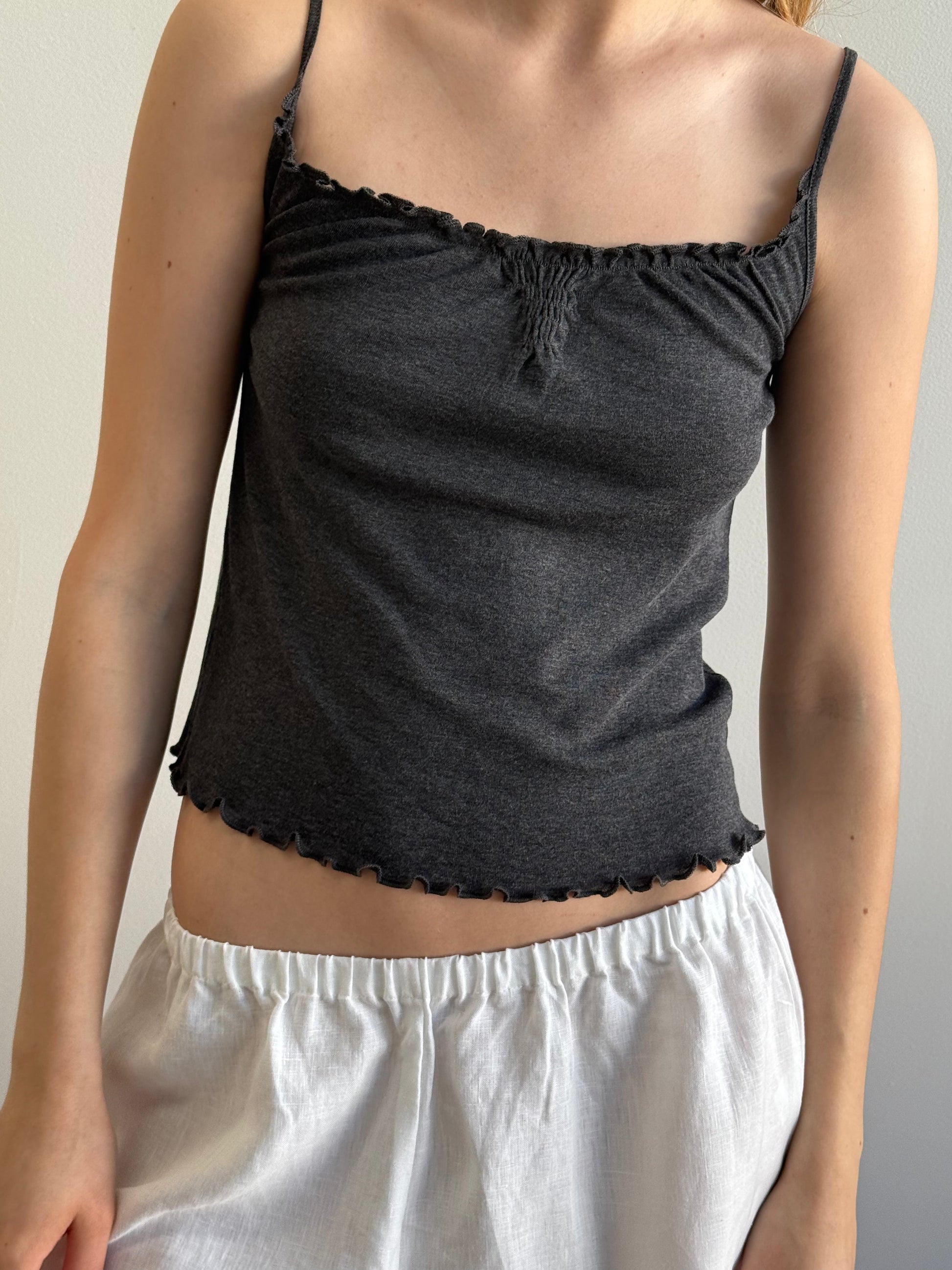 Wearing the Flounce Tank - Grey from Deiji Studios, a person pairs this dark grey, sleeveless top with ruffled edges and loose white shorts while standing against a plain light background.