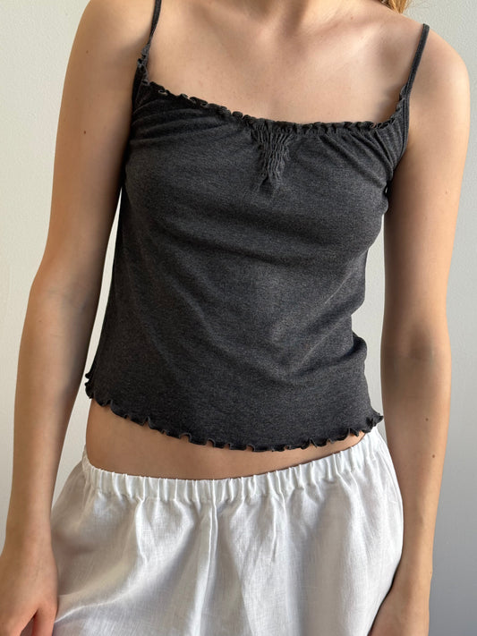 A person wearing the Wool Blend Flounce Tank - Grey with thin straps and ruffled edges, paired with loose white shorts, stands against a plain light background. Their face is not visible.