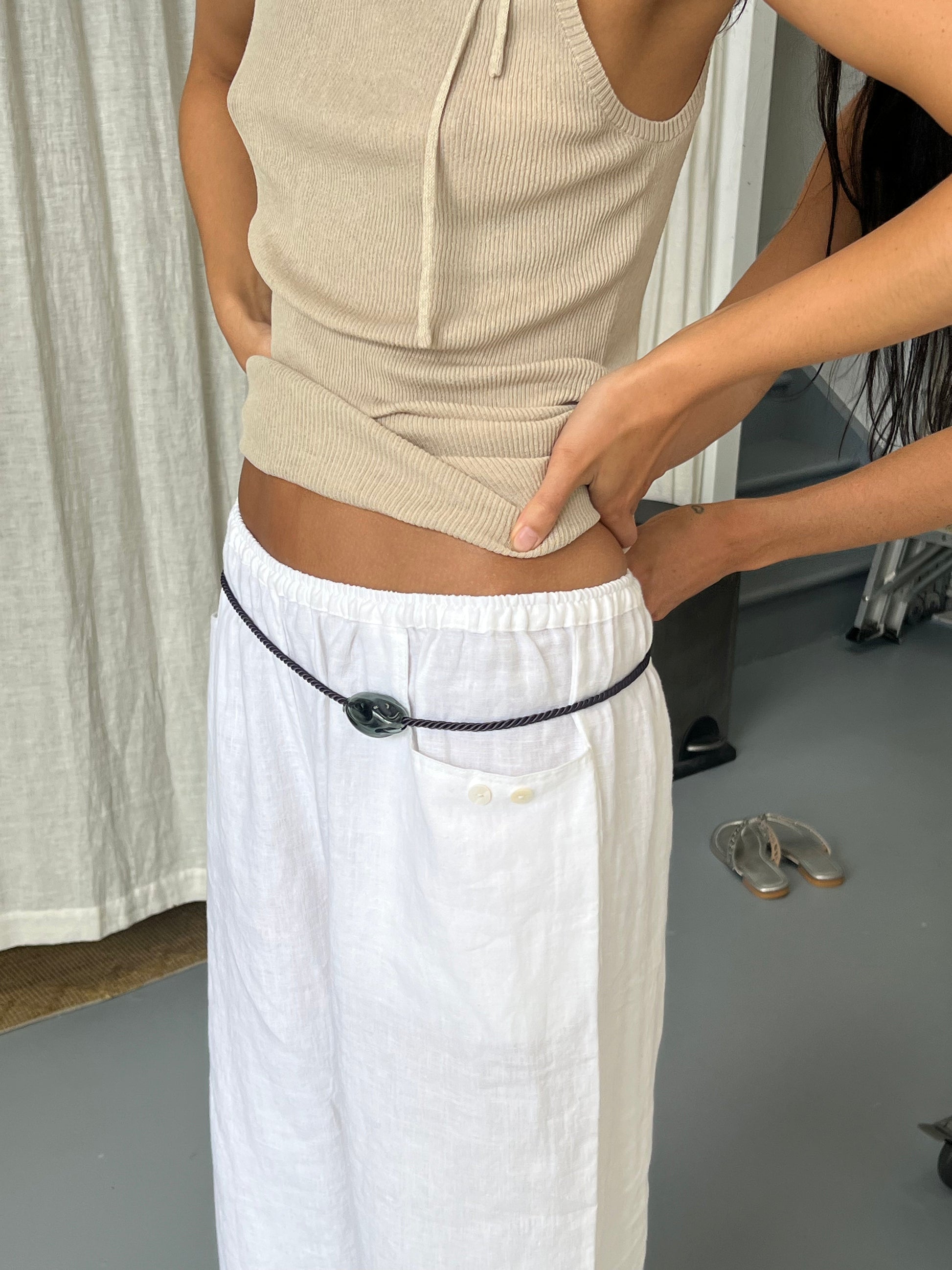 A person wears the Linen Square Pocket Pant - White by Deiji Studios with a beige sleeveless top, cinched by a thin belt with a large stone buckle. Another person adjusts the top while shoes and curtains appear in the background.
