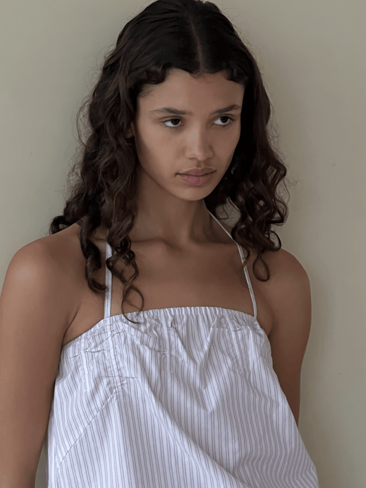A young woman with wavy brown hair wears the Tube Top - Musk Stripe by 0, featuring thin straps and a pinstriped cropped fit, standing against a plain light-colored wall with a neutral expression.