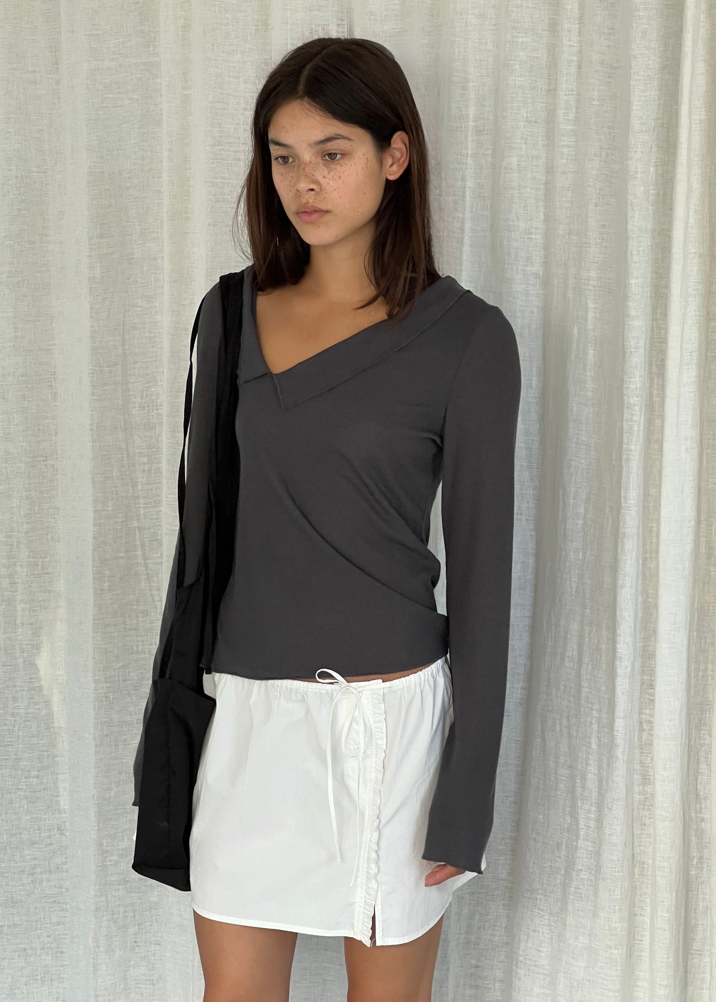A woman with straight dark hair stands before white curtains, wearing a Deiji Studios Cross Collar Top in night blue, a short white drawstring skirt, and carrying a black shoulder bag.