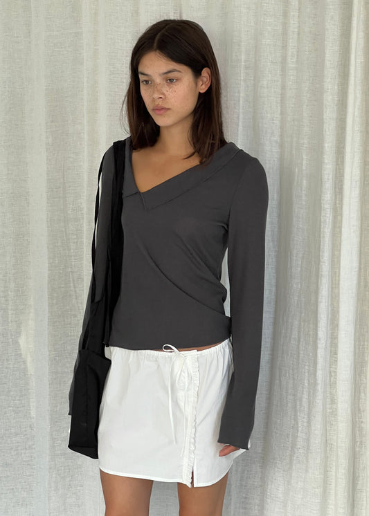 A woman with straight dark hair stands before white curtains, wearing a Deiji Studios Cross Collar Top in night blue, a short white drawstring skirt, and carrying a black shoulder bag.