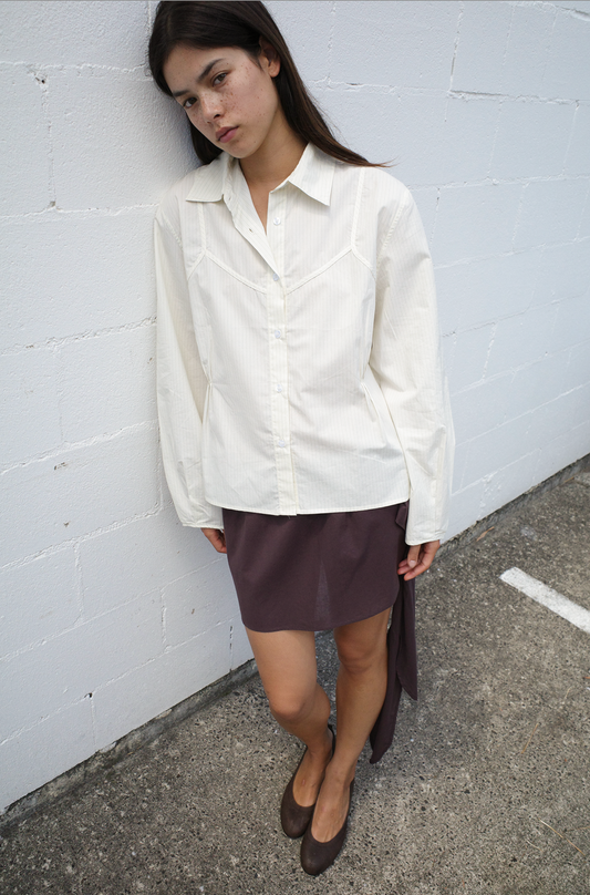 A woman with long brown hair leans against a white brick wall, wearing the Cami Overshirt – Citrus Stripe by Deiji Studios with a brown asymmetrical skirt and shoes, looking thoughtfully at the camera.
