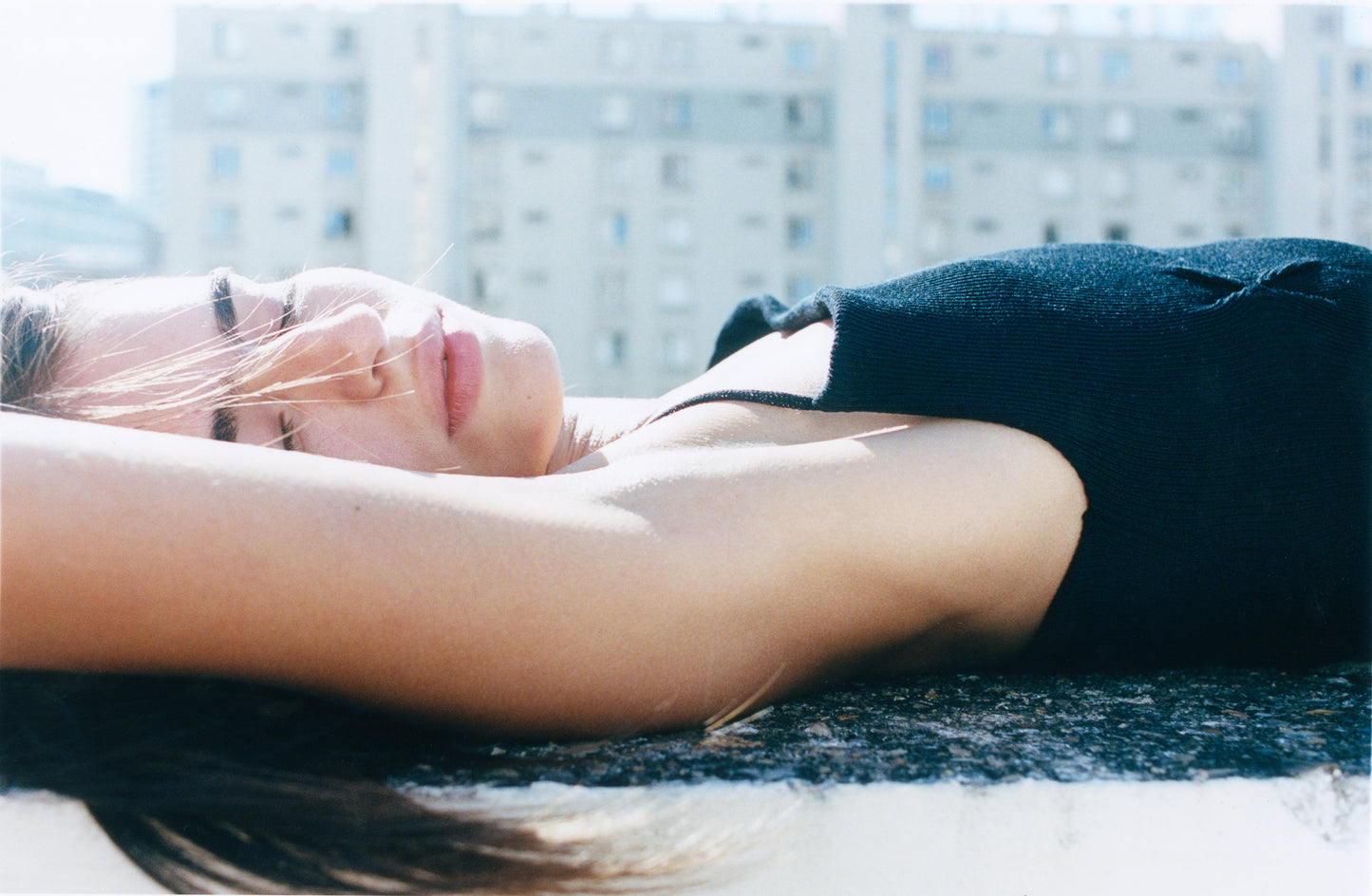 A person wearing the Fine Knot Tank - Black lies on their back outdoors, eyes closed and basking in sunlight, with blurred buildings in the background.