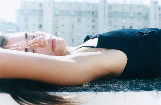 A person wearing the Fine Knot Tank - Black lies on their back outdoors, eyes closed and basking in sunlight, with blurred buildings in the background.