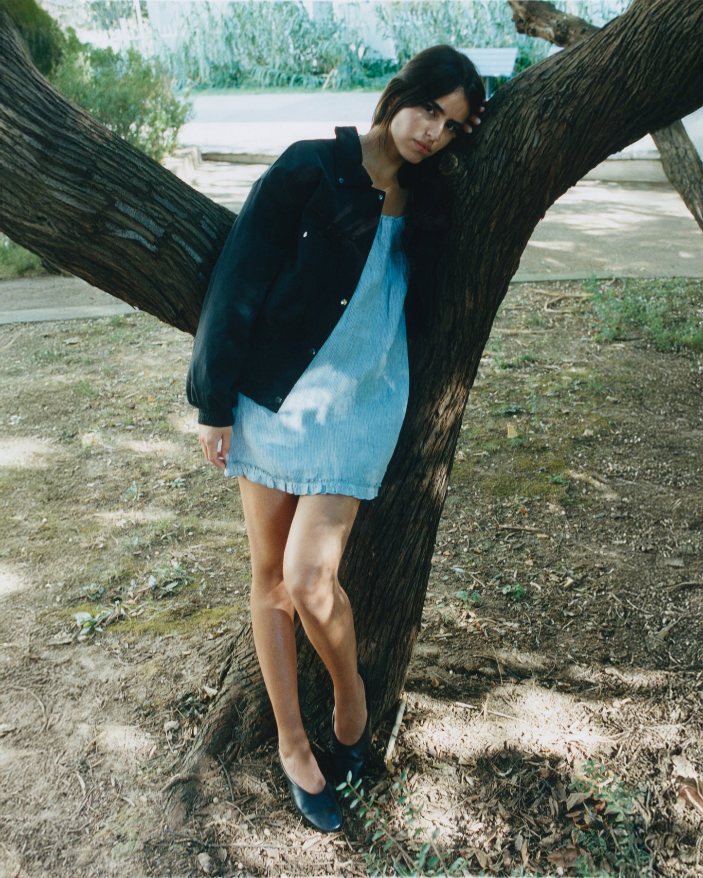 A woman in a blue dress and the Shirring Jacket - Slate by Deiji Studios leans against a tree in a park, standing on leaf-covered ground with one foot crossed over the other, her long hair falling as she looks slightly downward.