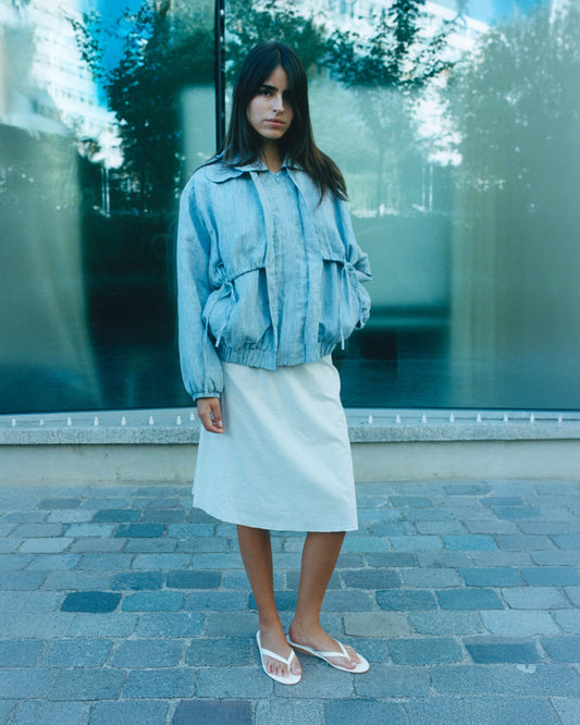 A woman stands outdoors on stone pavement, wearing the Double Layered Jacket - Blue Stripe over a knee-length light skirt and white sandals. A glass window reflecting trees is behind her.