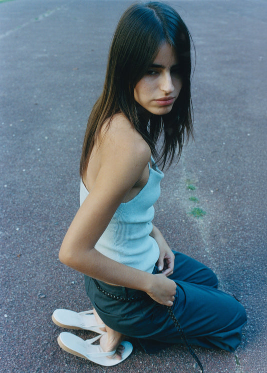 A young woman with long brown hair sits outdoors on a textured surface, wearing a light blue Deiji Studios tank top, Zip Pant - Charcoal, and white sandals, gazing to the side with a neutral expression.