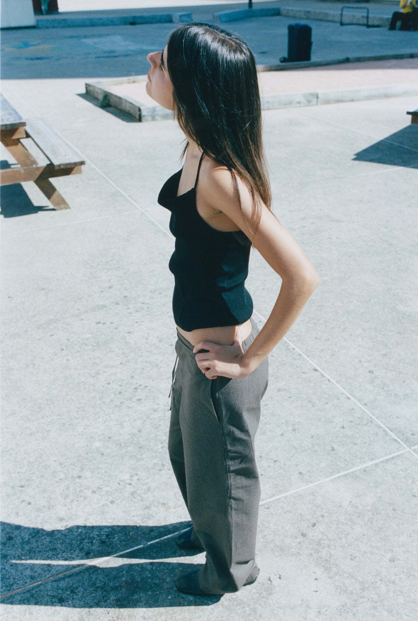 A young woman with long dark hair stands in profile on a sunny concrete plaza, wearing the Fine Knot Tank and loose grey trousers, hand on hip, gazing upward.