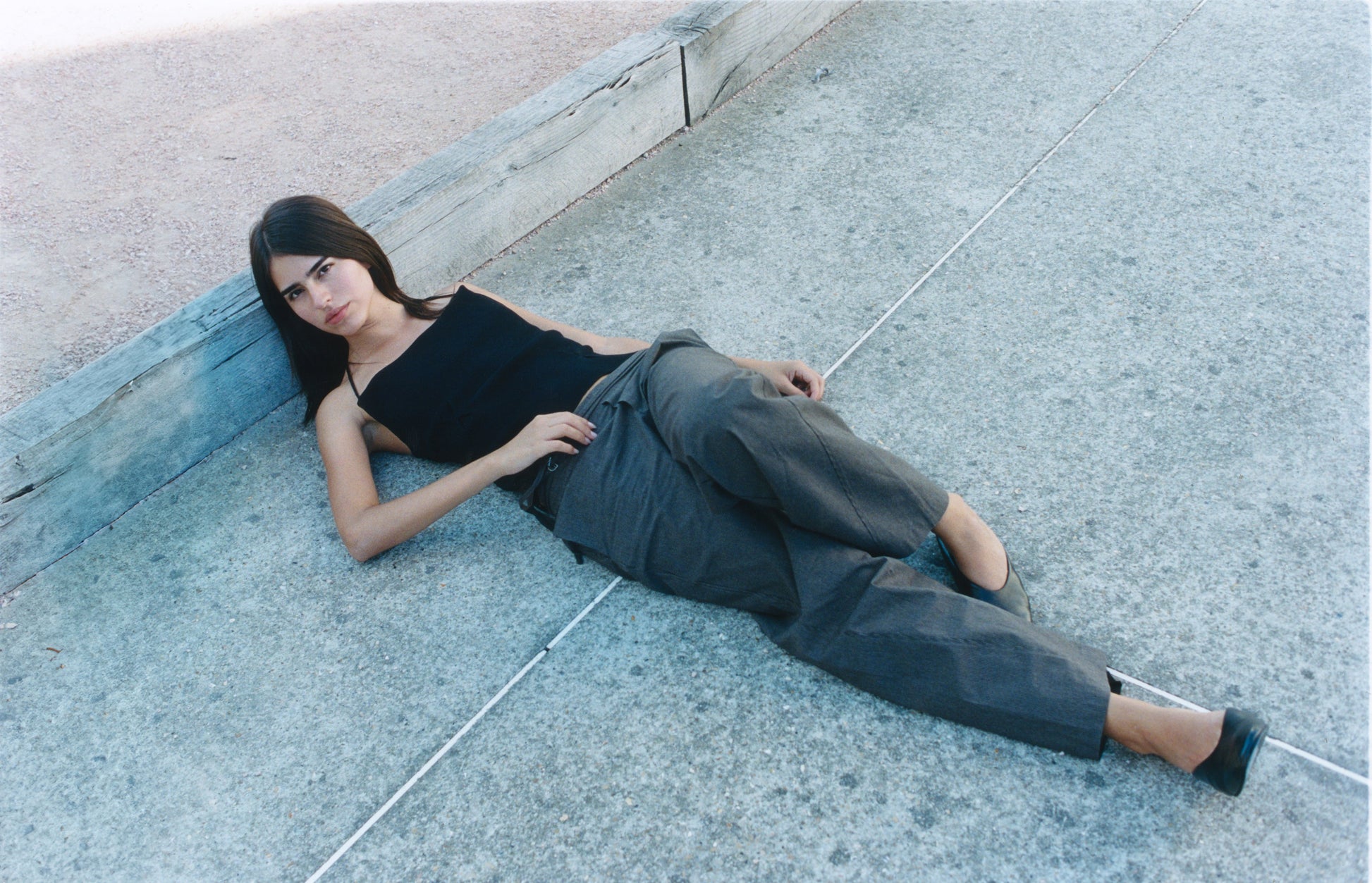 A woman in a Fine Knot Tank - Black and loose gray pants reclines on concrete, leaning against a wooden ledge with a relaxed, camera-facing gaze.