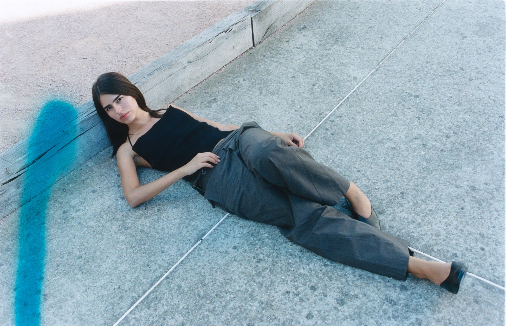 A woman with long dark hair, wearing a black tank top and the Double Belted Pant - Brown Stripe, lies on a concrete surface near a wooden beam. A blue streak is visible on the left side of the photo.
