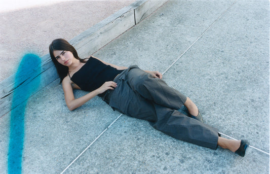 A woman with long dark hair, wearing a black tank top and the Double Belted Pant - Brown Stripe, lies on a concrete surface near a wooden beam. A blue streak is visible on the left side of the photo.