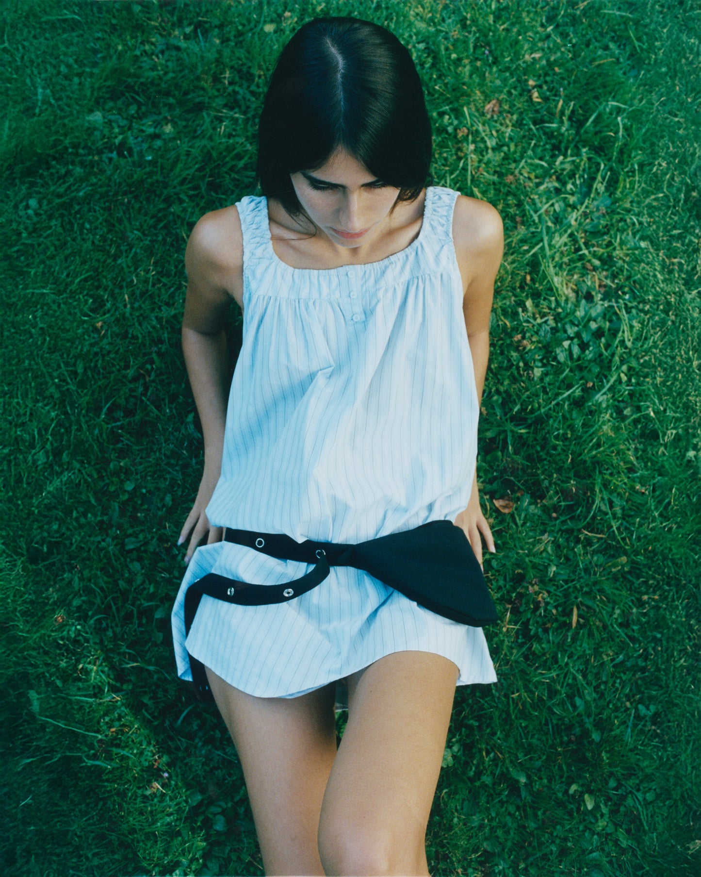 A woman with dark hair sits on grass outdoors, wearing a loose sleeveless light blue dress with a black sweater tied at her waist and the Snap Belt Bag - Black. The photo is shot from above in natural light.