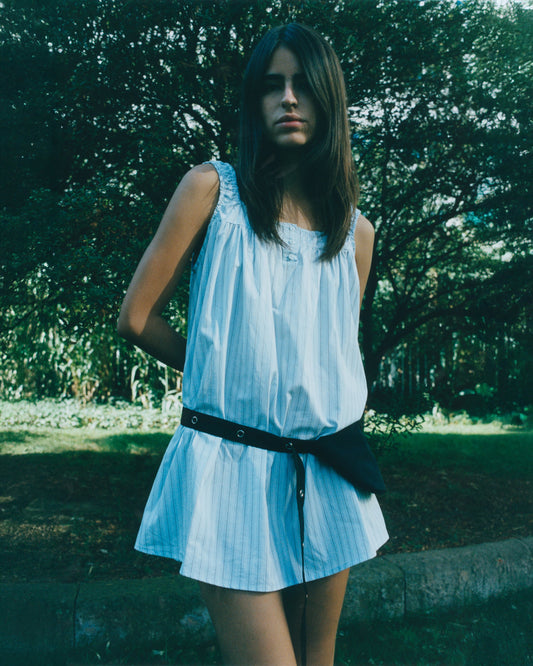 A woman with straight brown hair stands outdoors in front of greenery, wearing a sleeveless white dress with blue stripes and the Snap Belt Bag - Black at her waist, her hands behind her back.