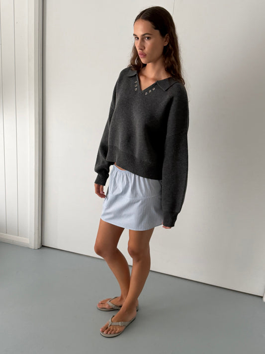 A young woman with long brown hair and a neutral expression stands indoors, wearing the Deiji Studios Eyelet Knit - Dark Pebble sweater along with a light blue skirt and beige sandals, posed against a plain white wall and light floor.