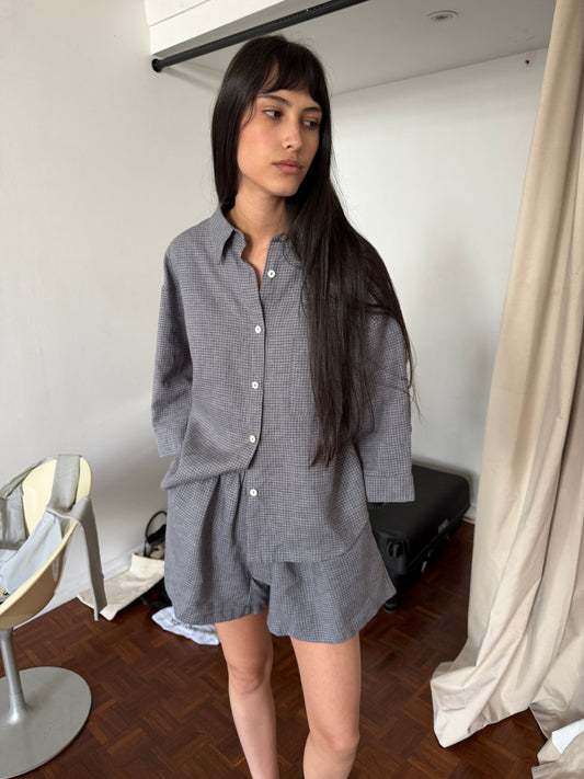 A cropped side view of the 03 Set - Indigo Check by Deiji Studios, showing the oversized boxy check linen shirt and matching shorts, worn by a woman with long dark hair in a room with a parquet floor.