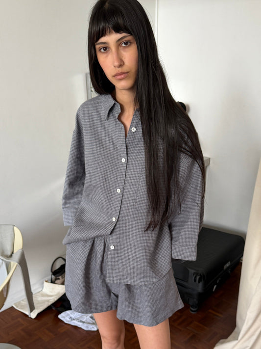 A woman with long dark hair and a fringe stands in a room with one hand in her pocket, wearing the 03 Set - Indigo Check by Deiji Studios, an oversized boxy check linen shirt with matching boxer-style shorts.
