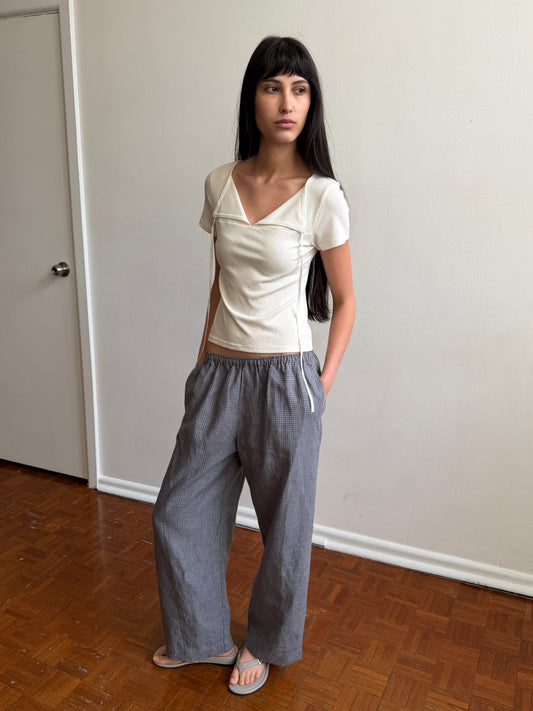 A woman with dark hair and a fringe stands against a door wearing the Tie Back Tee - White by Deiji Studios, a fitted cream tee with a sweetheart neckline, with grey check trousers and flip-flops.