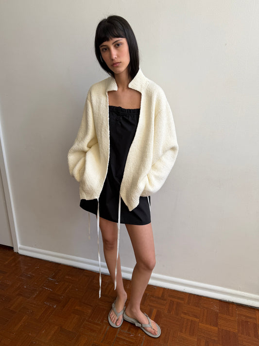 A woman with dark hair and a fringe stands against a wall wearing the Wrap Cardigan - Snow by Deiji Studios, an oversized cream knit wrap cardigan with a raised collar, over a black top and mini skirt with flip-flops.