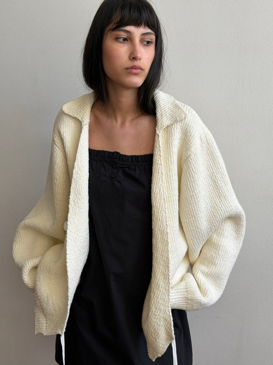 A woman with dark hair and a fringe wears the Wrap Cardigan - Snow by Deiji Studios, an oversized cream knit wrap cardigan with a collar, open over a black top.