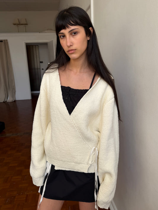 A woman with dark hair and a fringe stands in a room wearing the Wrap Cardigan - Snow by Deiji Studios, a cream knit wrap cardigan with a collar and waist tie, over a black top and mini skirt.