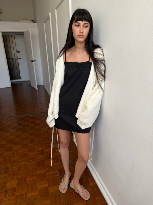 A woman with dark hair and a fringe leans against a wall wearing the Wrap Cardigan - Snow by Deiji Studios, an oversized cream knit wrap cardigan, over a black slip dress with flip-flops.