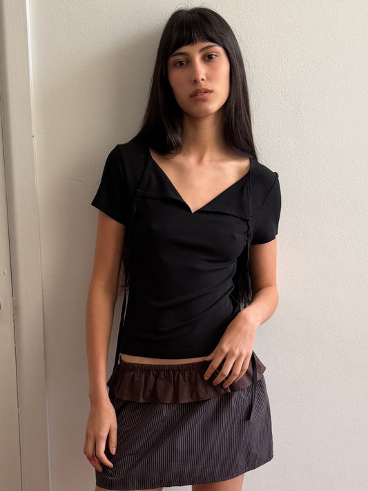 A woman with dark hair and a fringe leans against a wall wearing the Tie Back Tee - Black by Deiji Studios, a fitted black tee with a sweetheart neckline and back ties, with a brown flounce mini skirt and flip-flops.