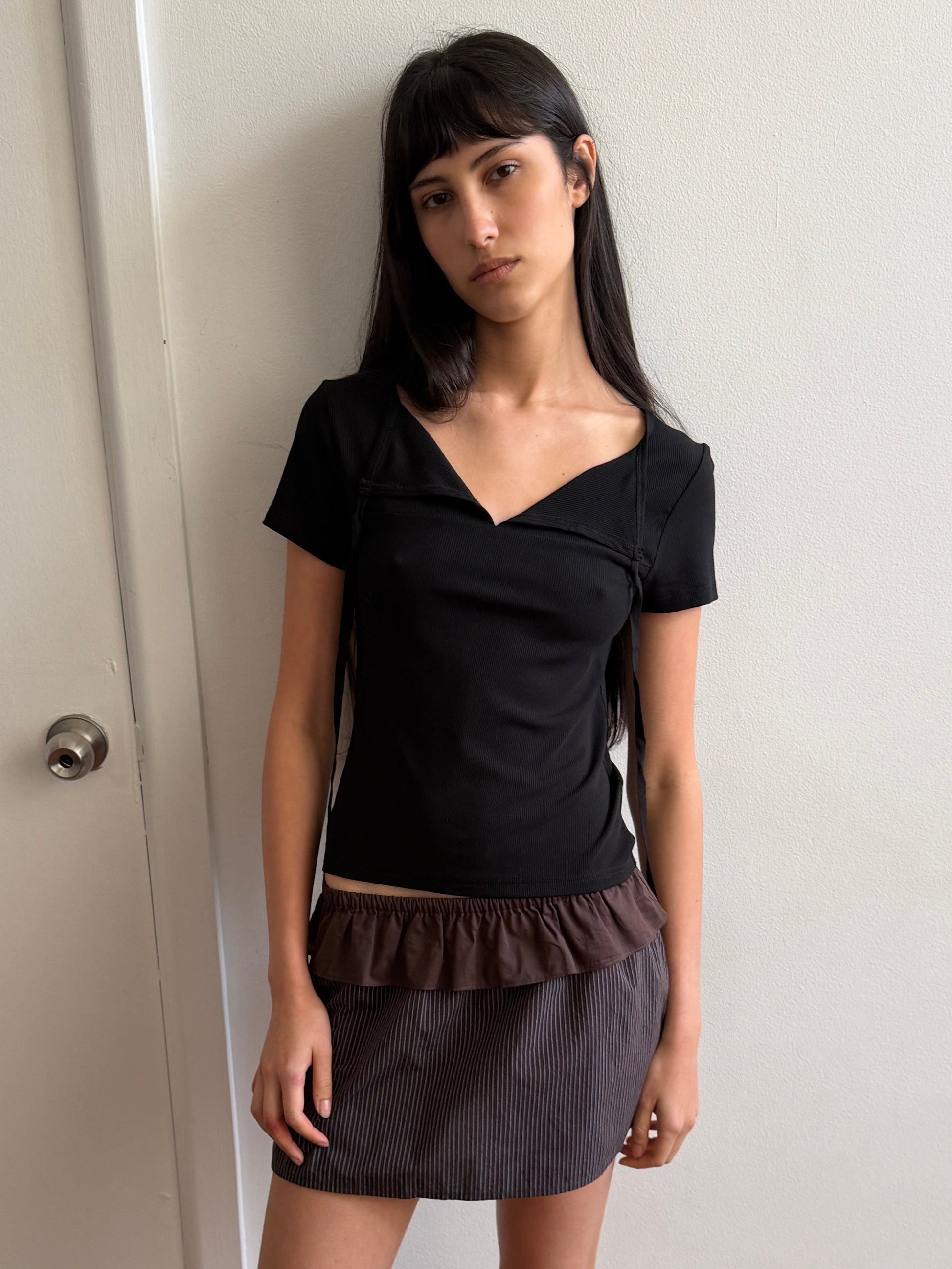 A woman with dark hair and a fringe stands against a door wearing the Tie Back Tee - Black by Deiji Studios, a fitted black tee with a sweetheart neckline, with a brown flounce mini skirt and flip-flops.