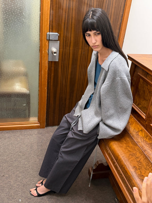 A woman with dark hair and a fringe sits on a wooden bench wearing the Wrap Cardigan - Grey by Deiji Studios, an oversized grey knit wrap cardigan, over a blue top with pinstripe trousers.