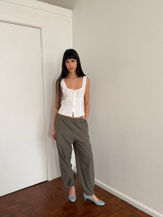 A woman with dark hair and a fringe stands in a room wearing the Hem Toggle Pant - Moss Stripe by Deiji Studios, moss-green trousers with a toggle-gathered hem, with a white tie top and pale blue heels.