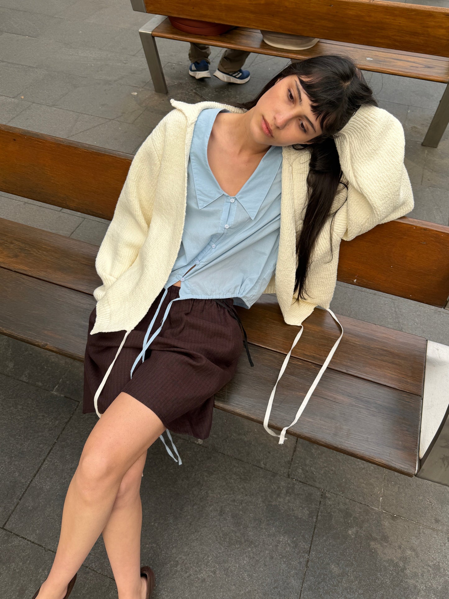 A woman with long dark hair reclines on an outdoor bench wearing the Shoulder Tucks Top - Baby Blue by Deiji Studios, a baby blue blouse, under a cream cardigan with brown shorts.