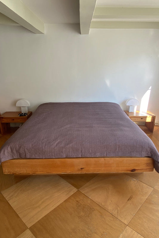 A minimalist bedroom features a wooden platform bed with a Russet Check US duvet cover, two wooden nightstands with white lamps, light brown tile flooring, plain white walls, and exposed ceiling beams.