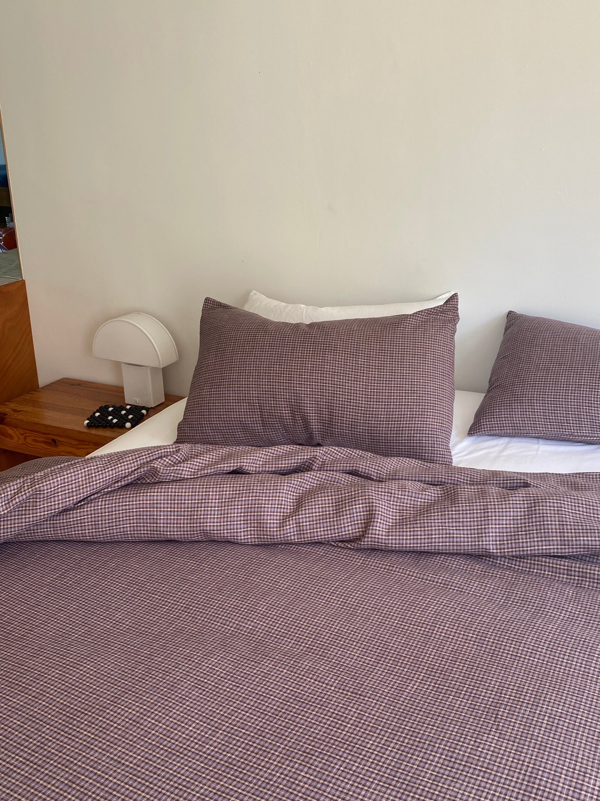 A neatly made bed with purple checkered bedding, a Pillow slip set - Russet Check from Deiji Studios, and a white headboard beside a small wooden nightstand with a white lamp and a polka-dotted eye mask.