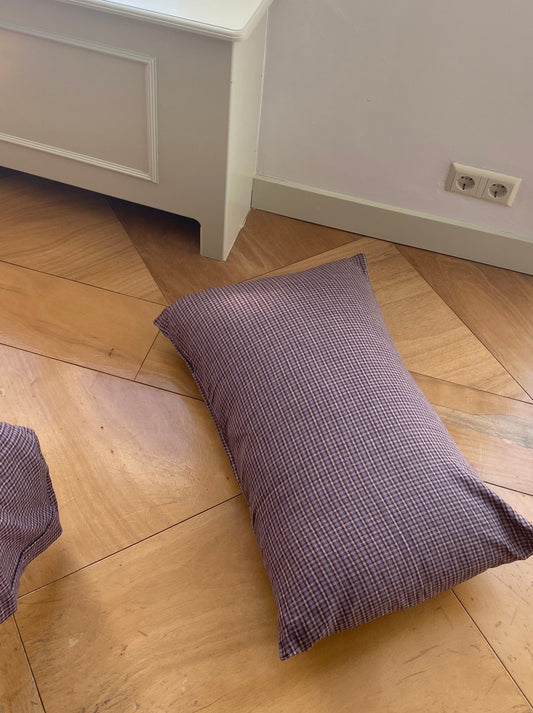 A Pillow Slips rectangular checkered pillow in purple and white rests on a wooden floor near white furniture and an electrical outlet.