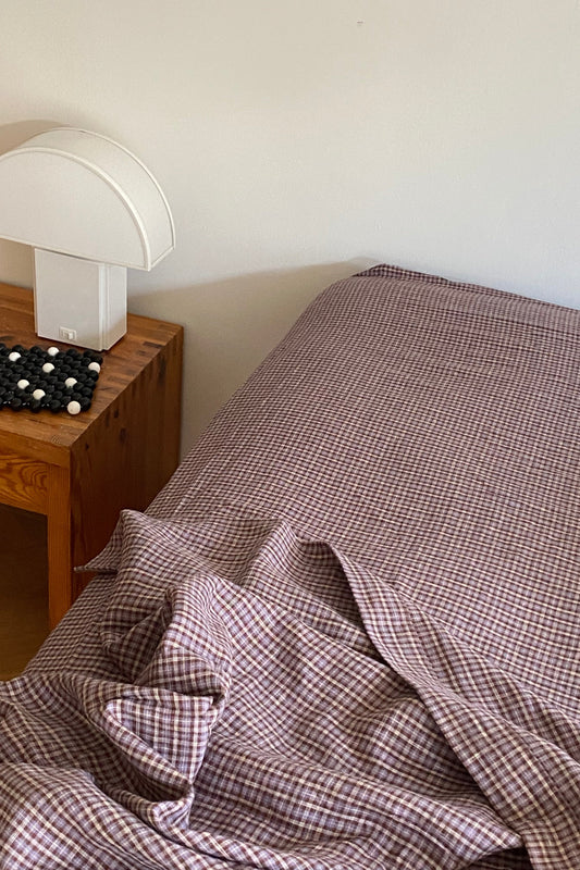 Brown and white checkered Sheets cover a bed beside a wooden nightstand topped with a white modern lamp and a decorative black and white beaded object.
