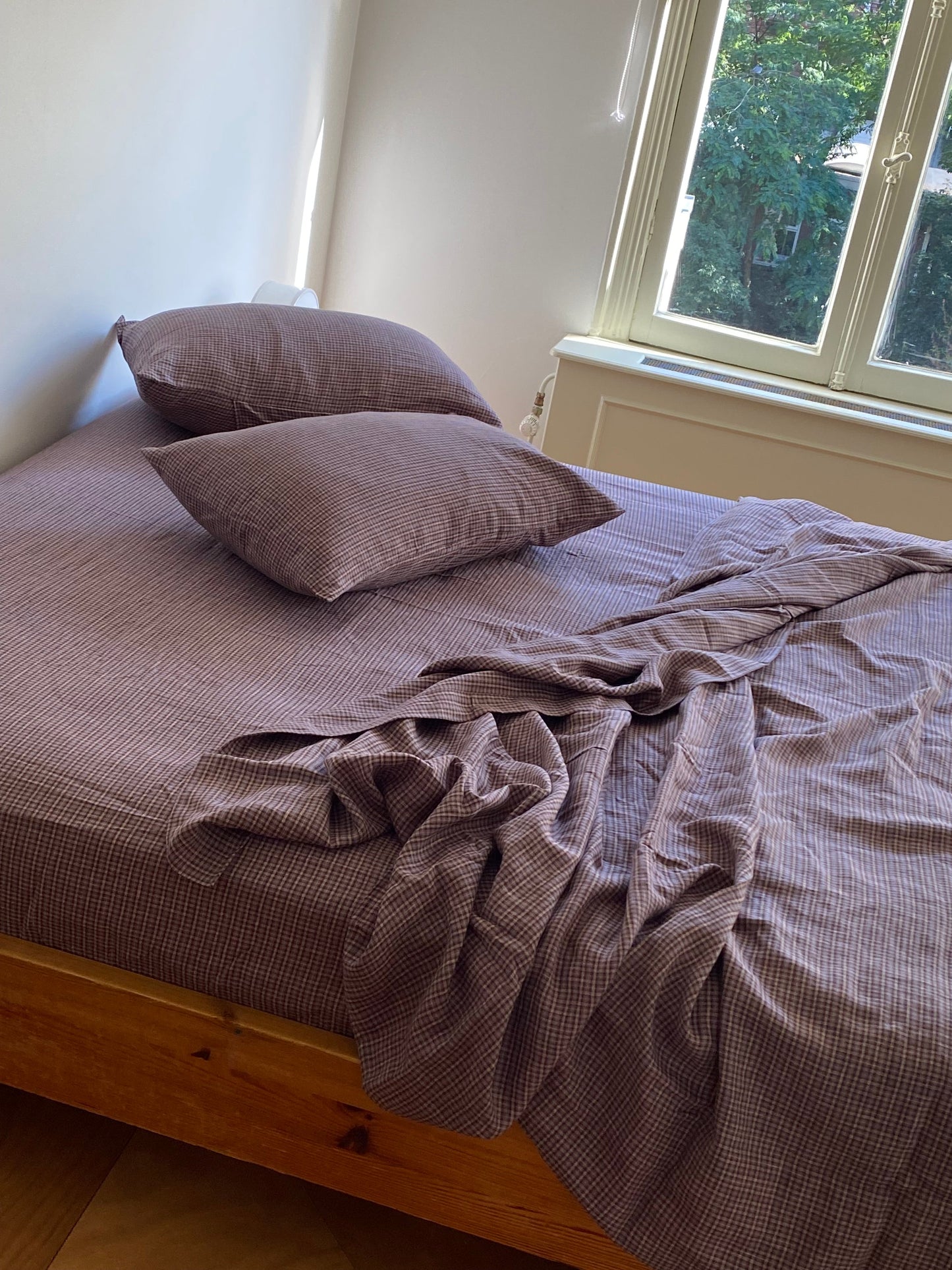 A wooden-framed bed with brown checkered sheets and two Pillow slip set - Russet Check slips by Deiji Studios sits near a sunlit window with trees outside; the bedding is slightly rumpled.