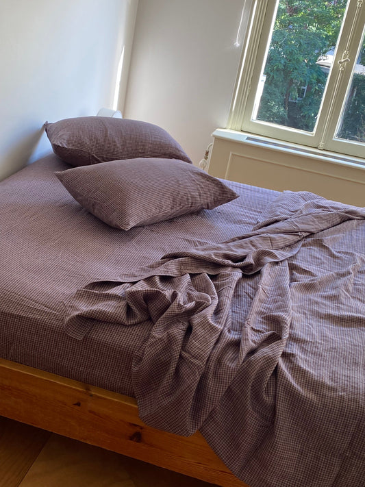 A wooden bed with brown-checkered sheets and two hypo-allergenic pillows in the Pillow slip set - Russet Check is placed by a sunlit window overlooking green trees.
